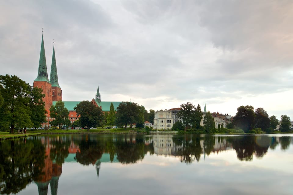 A calm lake reflecting a church with two spires and surrounding greenery.