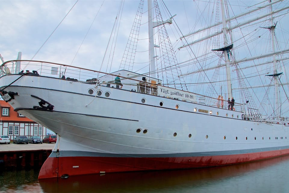 A large sailing ship docked at a harbor with a red hull and white superstructure.