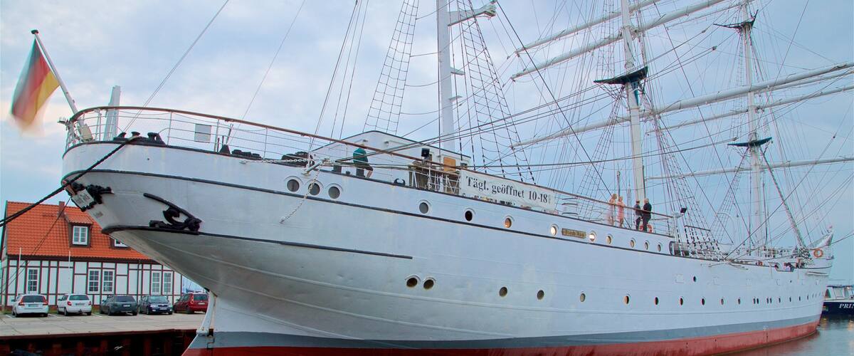A large sailing ship docked at a harbor with a red hull and white superstructure.