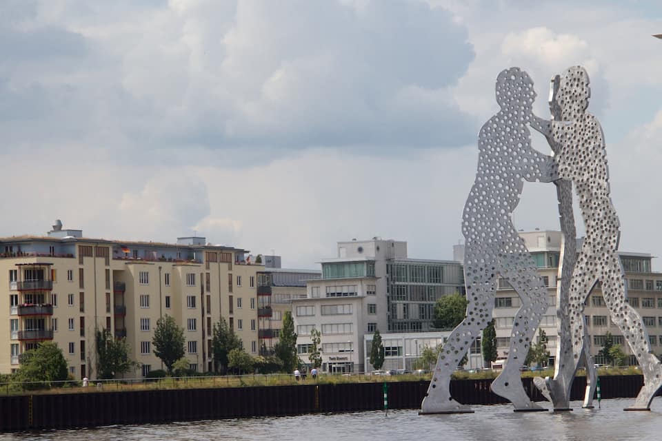 Escultura de dos figuras en una pose de baile frente a un muelle, con edificios modernos al fondo.