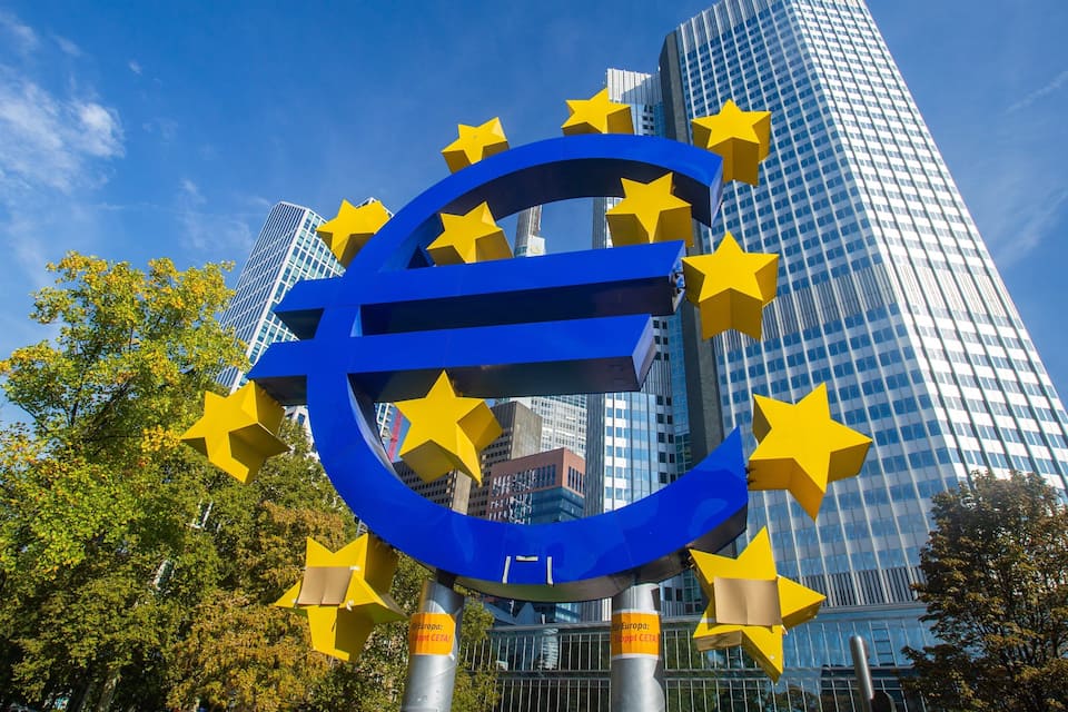 Um grande símbolo do euro em azul com estrelas amarelas, rodeado por edifícios altos e árvores.