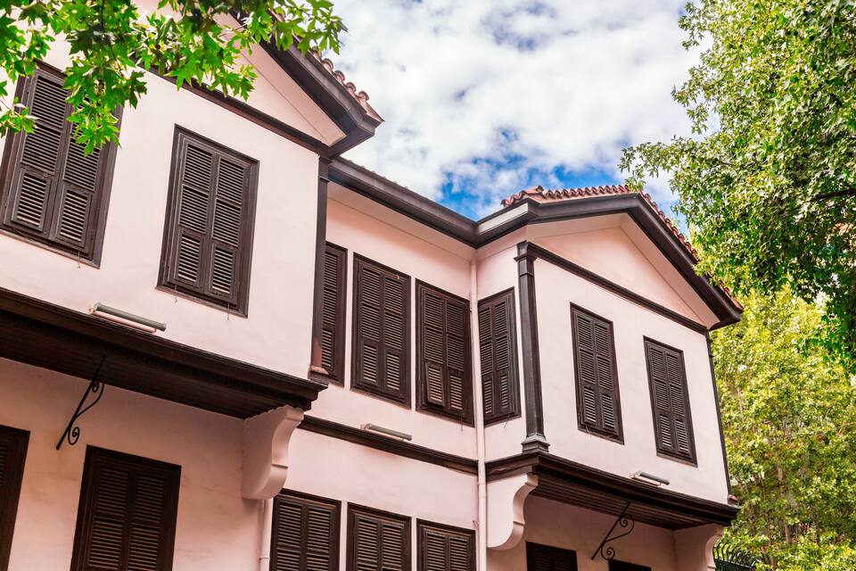 Ein pinkfarbenes Gebäude mit zahlreichen Fenstern und Fensterläden, umgeben von Bäumen.