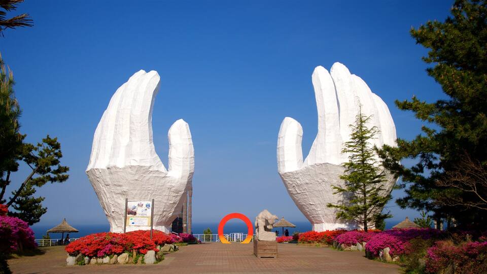 A large white sculpture of hands in a garden with red flowers and a clear blue sky.