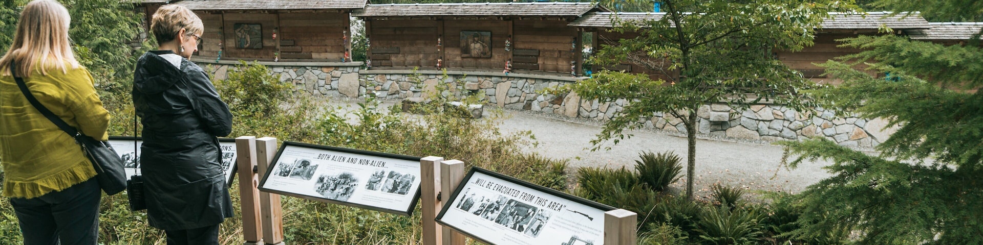 Duas pessoas em frente a um sinal de madeira com fotos históricas, com vista para uma cabana rústica e uma floresta densa.