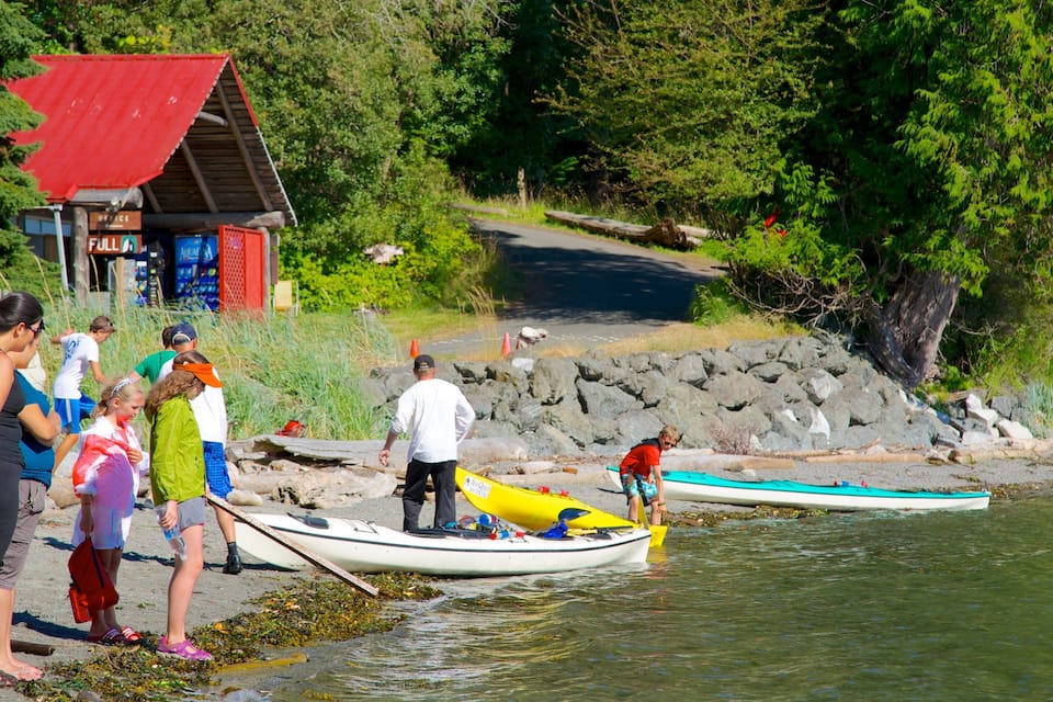 Personas preparando kayaks en la orilla de un río, con una pequeña oficina y un edificio de techo rojo al fondo.