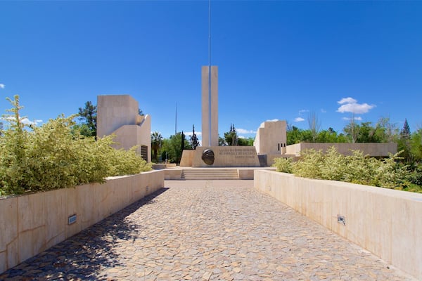 A monument with a tall central pillar and a plaque with text, surrounded by a paved pathway and greenery.