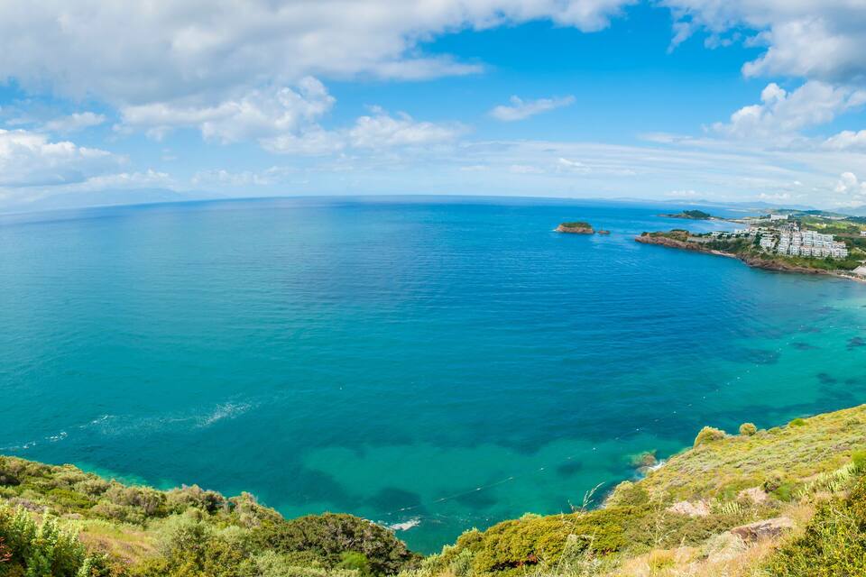 A coastal landscape with clear turquoise waters, a small island, and a building complex on the shore.