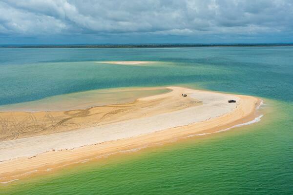 An aerial view of a coastal area with a sandy beach, clear turquoise waters, and distant land.