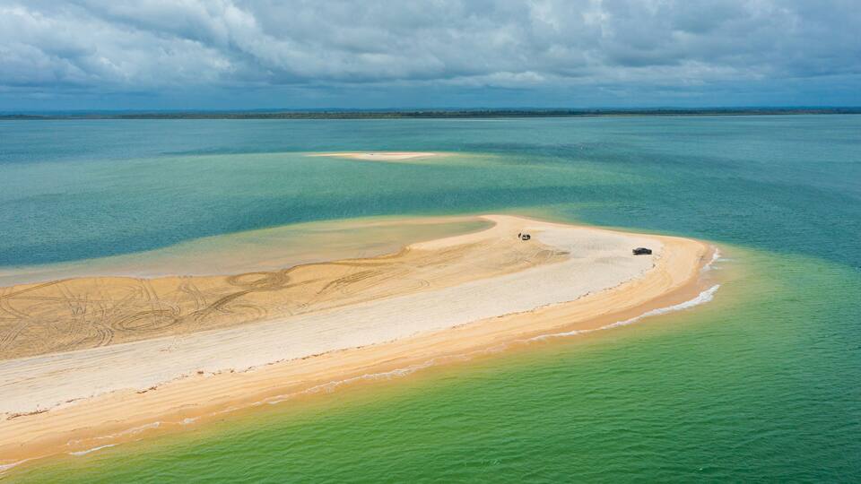 An aerial view of a coastal area with a sandy beach, clear turquoise waters, and distant land.