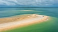 An aerial view of a coastal area with a sandy beach, clear turquoise waters, and distant land.