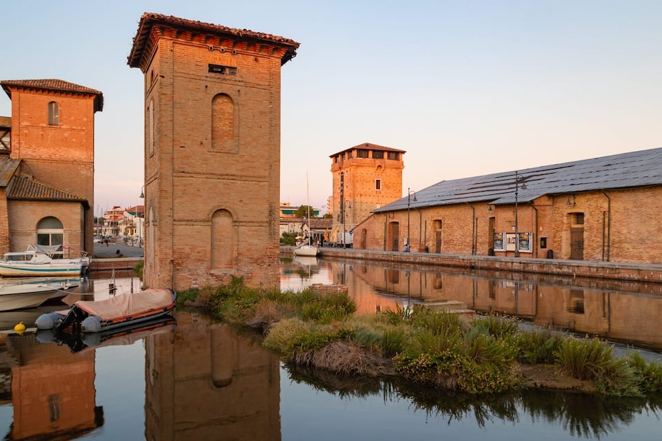 Un canale con imbarcazioni, una torre e edifici in mattoni riflessi nell'acqua.