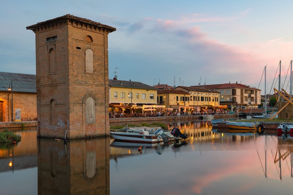 Una torre histórica, un canal con barcos y edificios frente al agua que se reflejan en ella.