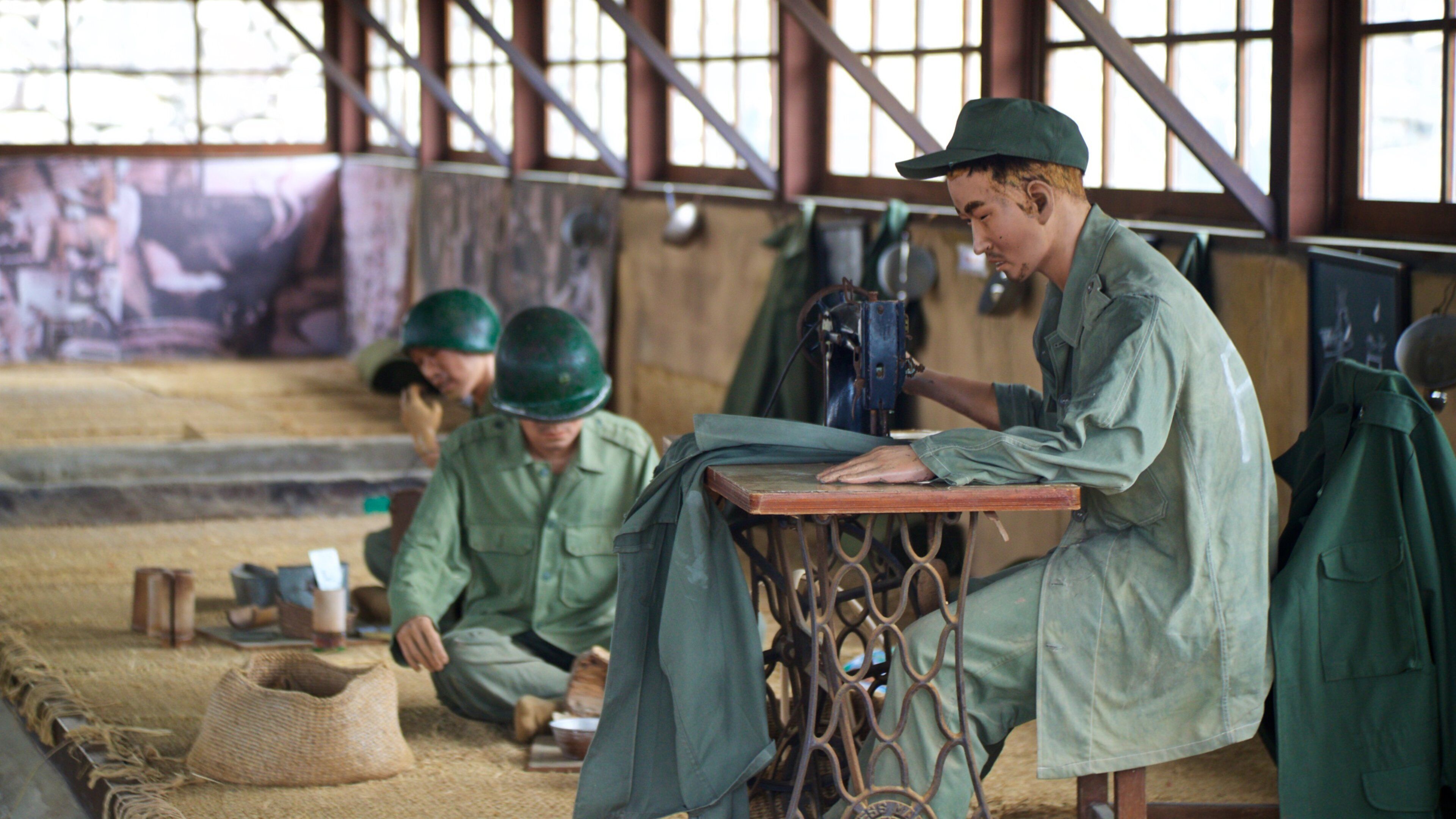 A workshop with workers in green uniforms and helmets, sewing machines, and a large window in the background.
