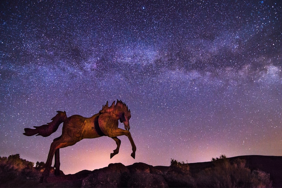 A starry night sky with the Milky Way visible, a large metallic sculpture of a horse, and a rocky landscape.