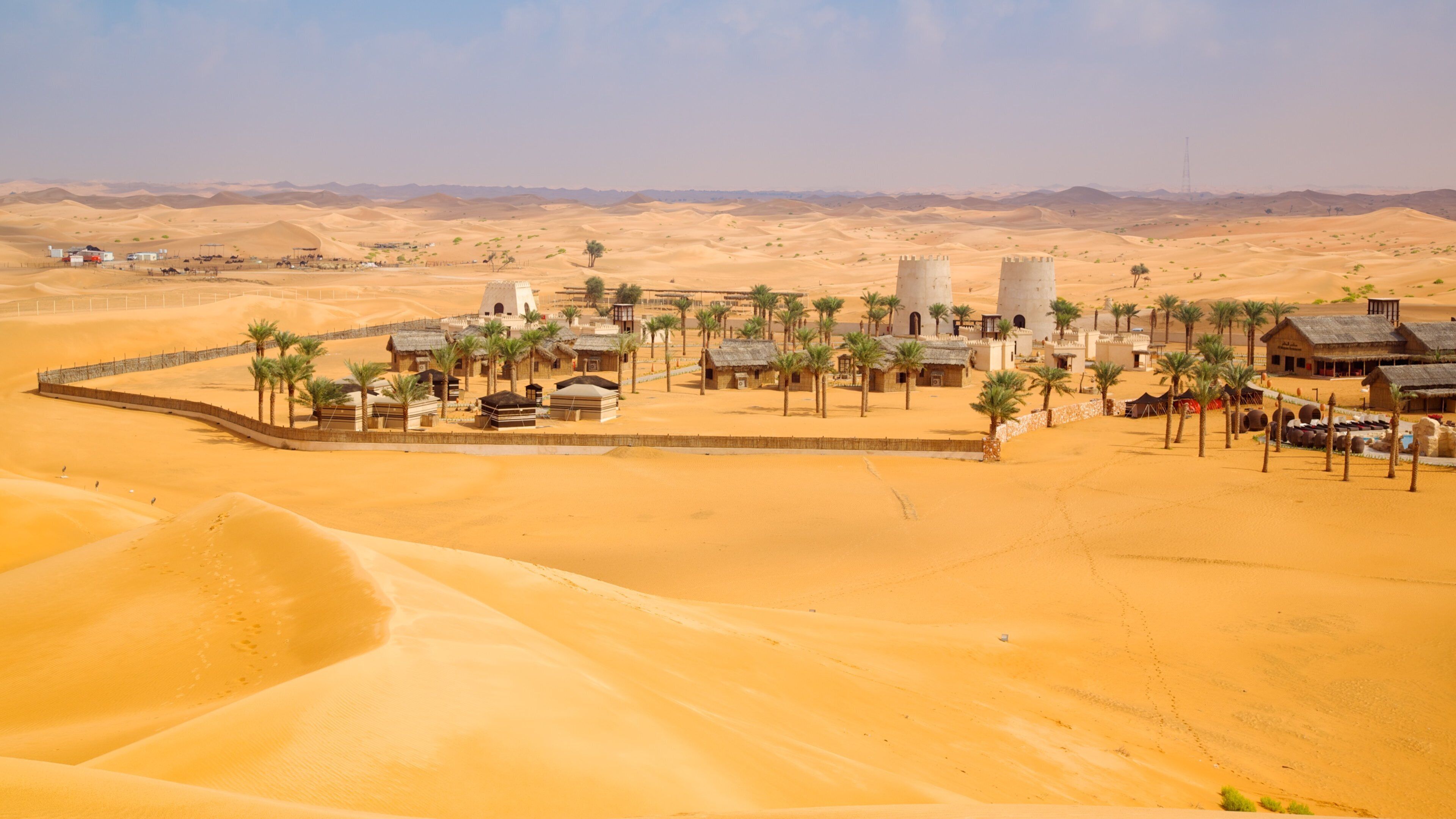 A desert landscape with sand dunes, a small settlement with palm trees, and a few buildings.