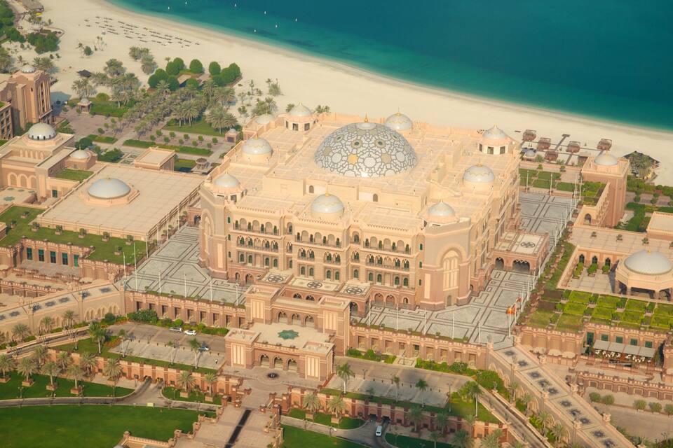 Une vue aérienne d’un grand bâtiment orné, doté de plusieurs dômes, entouré de palmiers et bordé par une plage.