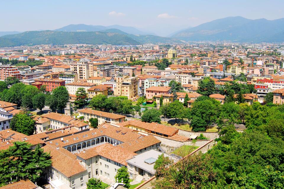 A cityscape with a mix of historic and modern buildings, surrounded by lush greenery and mountains in the background.