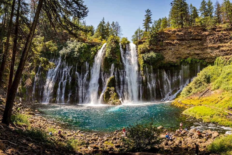 A multi-tiered waterfall cascading into a clear blue pool surrounded by forested cliffs.