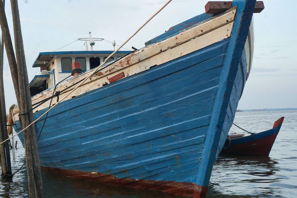Un bateau bleu avec une cabine, amarré à un quai grâce à des poteaux en bois.