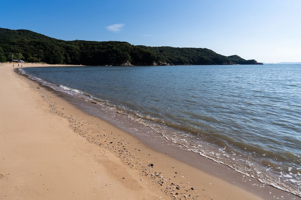 A sandy beach with gentle waves, a clear blue sky, and a forested hill in the background.