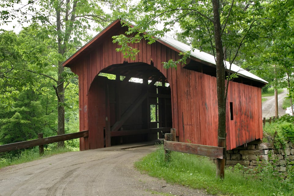 A red covered bridge in a wooded area with a dirt road leading to it.