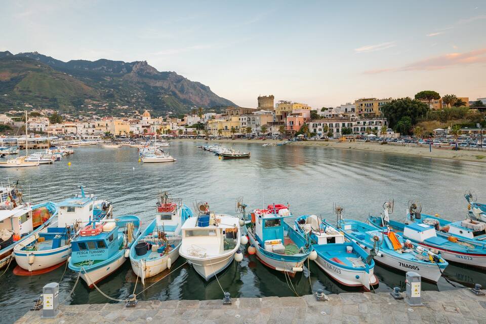 Un puerto con numerosos barcos amarrados, un telón de fondo montañoso y una ciudad costera con edificios.