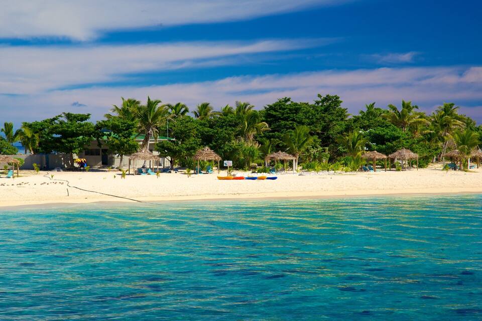 Een strand met helder turquoise water, palmbomen en hutten met rieten daken.