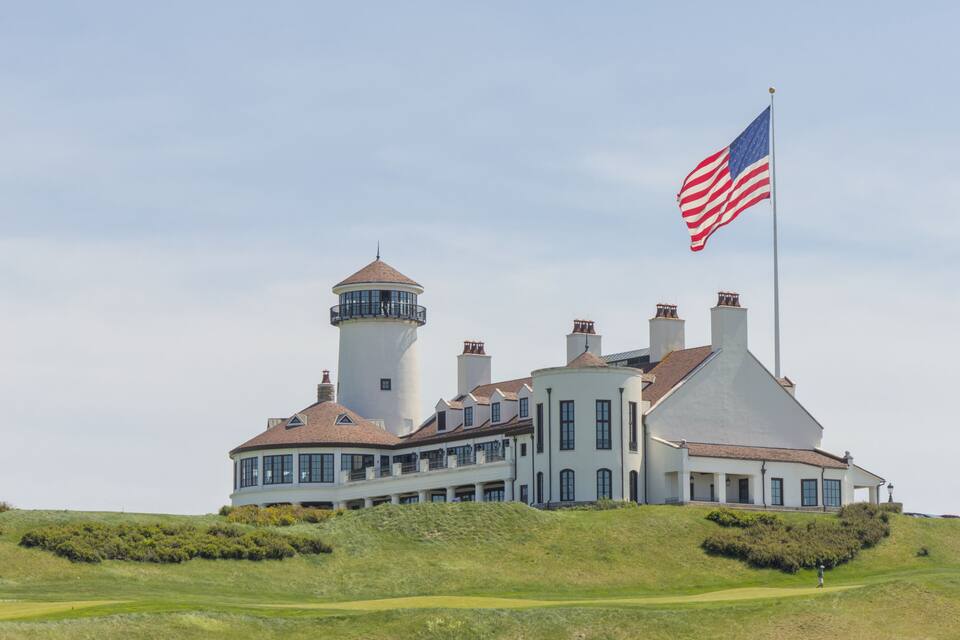Un grand bâtiment blanc surmonté d’une tour de phare, entouré de collines verdoyantes et arborant un drapeau américain.