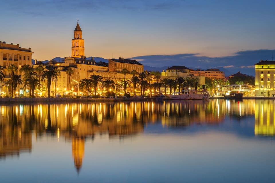 Paesaggio urbano sul lungomare con edifici illuminati, una torre imponente e palme riflesse in acque calme.