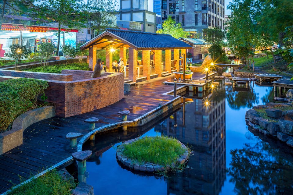 En rolig flodpromenade med træterrasse, en lille murbygning og en fredfyldt sø, der spejler omgivelserne.