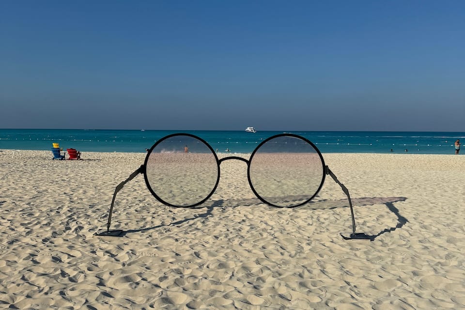 A pair of sunglasses on a sandy beach with a clear blue sky and a boat in the distance.
