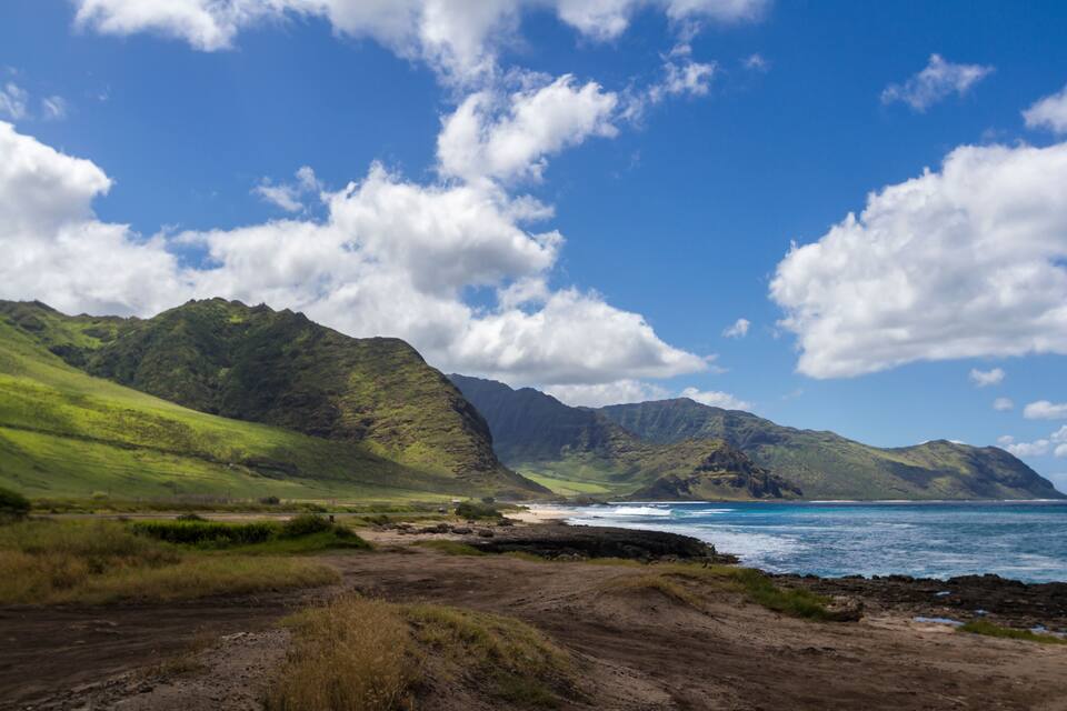 A coastal landscape with green hills, a beach, and the ocean under a blue sky with clouds.