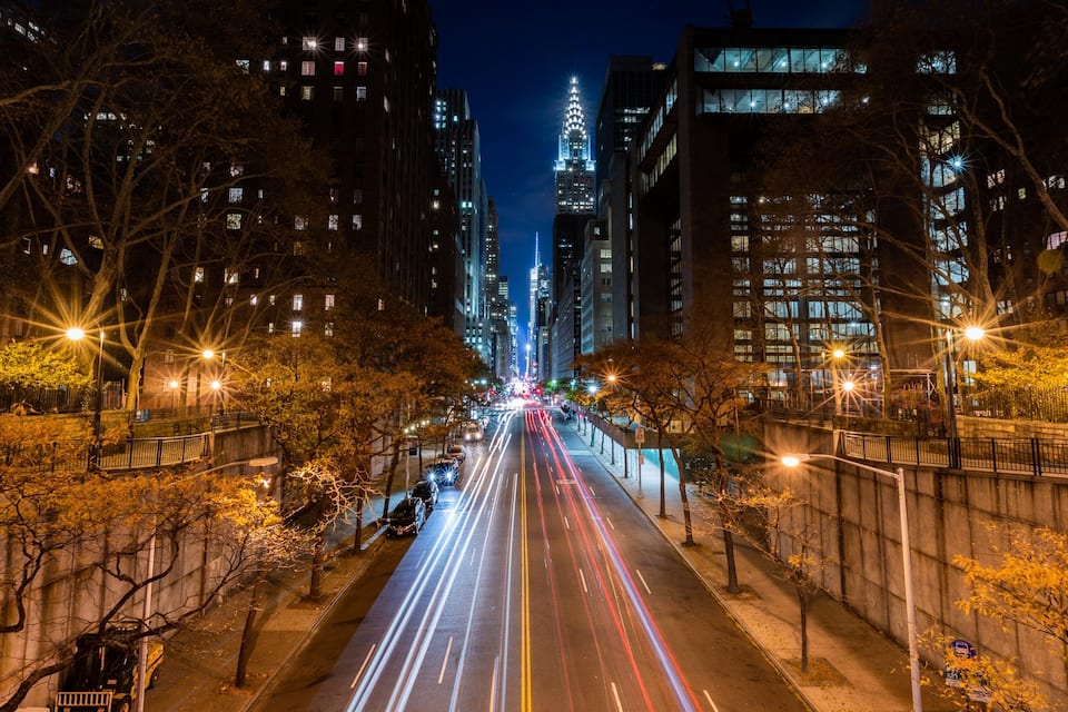 A nighttime cityscape with illuminated skyscrapers and a long exposure of light trails on the road.