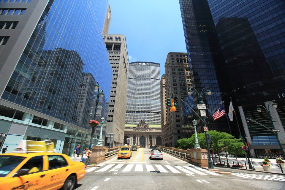Un encreuament urbà bullició envoltat d'rascos de cel moderns, incloent-hi un edifici MetLife, i un cel blau clar.
