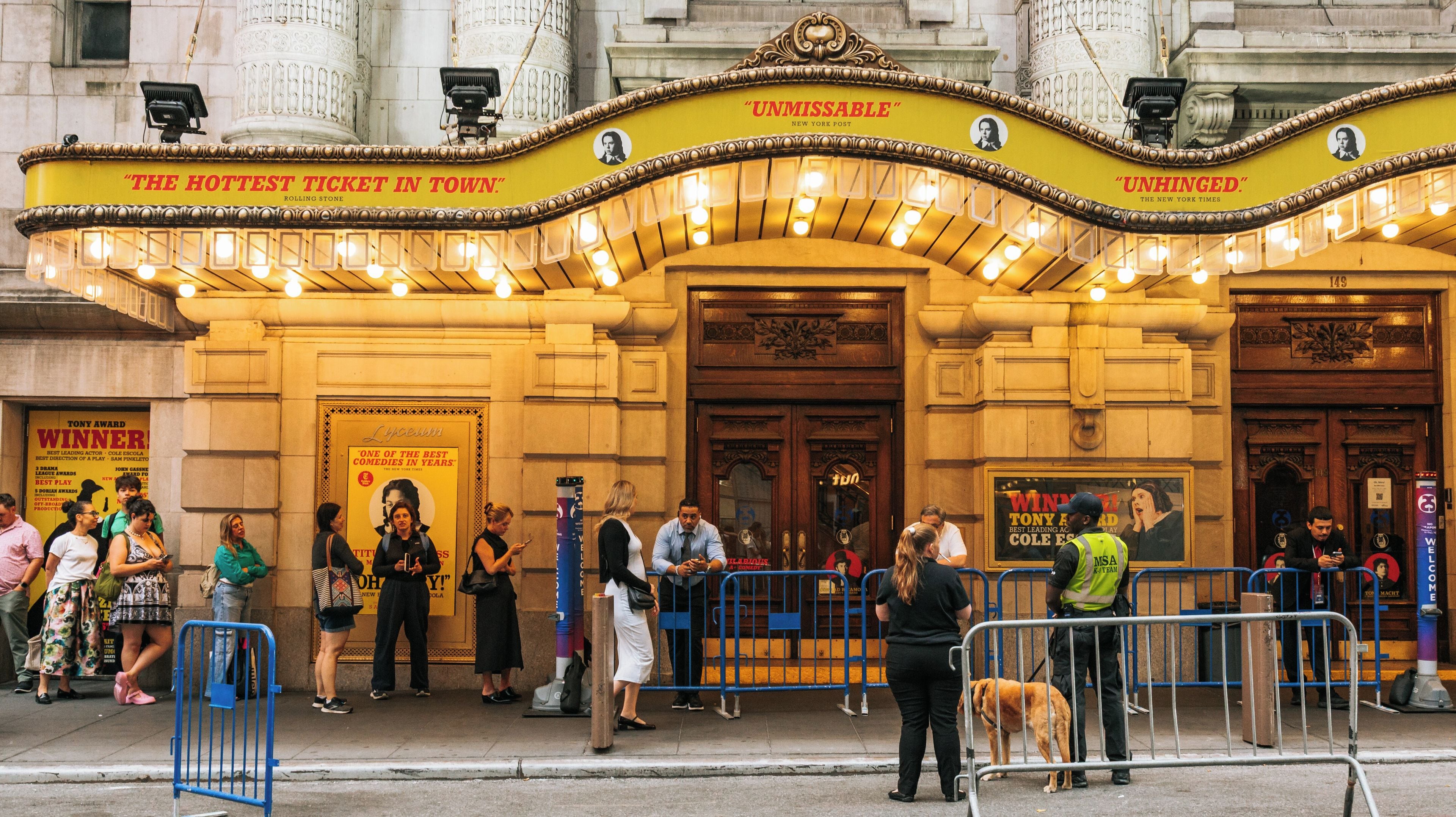 A theater marquee with 'The Hottest Ticket in Town' and 'Unmissable' signs, a crowd of people, and a dog on the sidewalk.