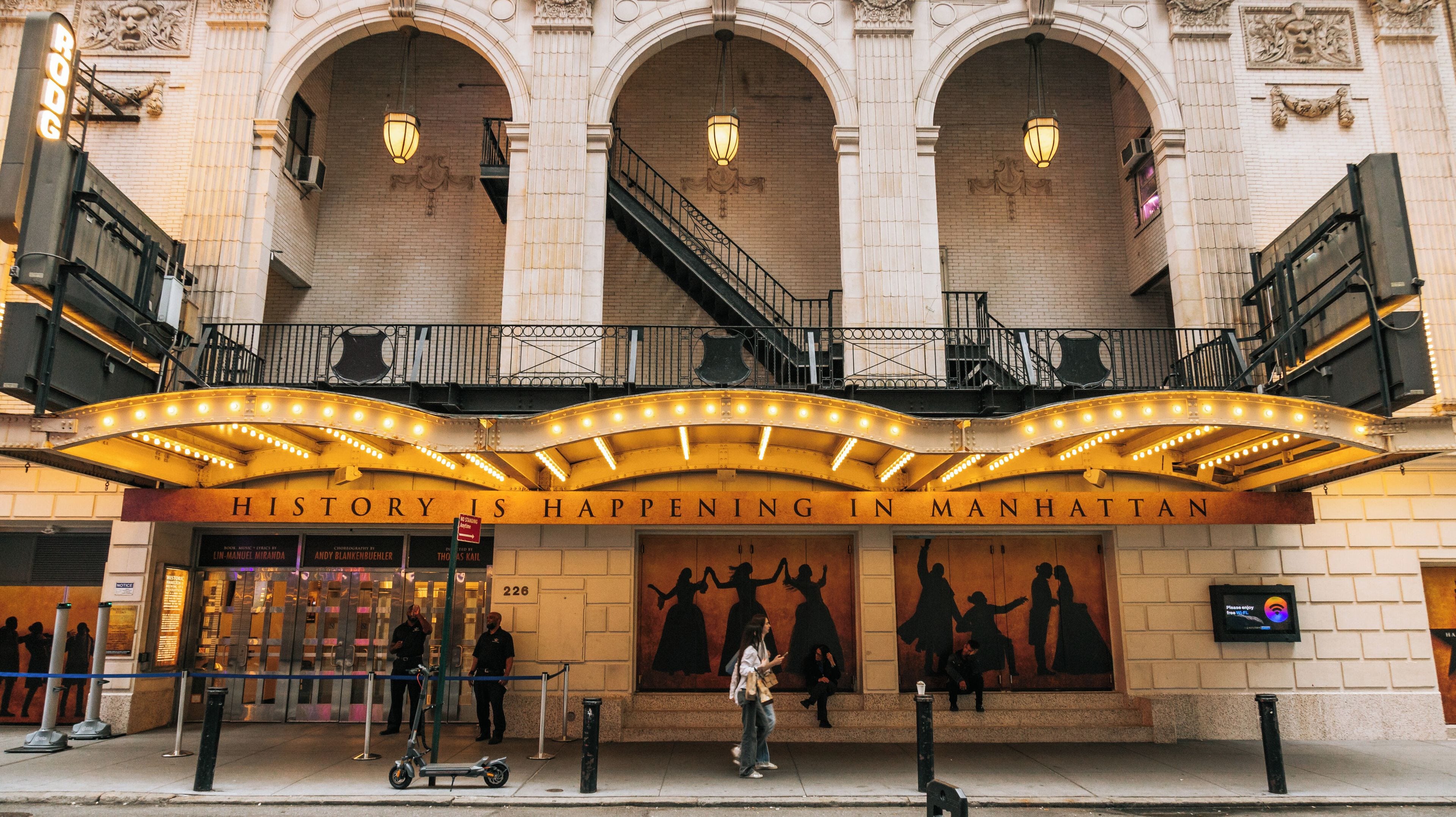 Un théâtre avec une enseigne lumineuse sur laquelle on peut lire : « L’HISTOIRE S’ÉCRIT À MANHATTAN », et un escalier menant à un niveau supérieur.