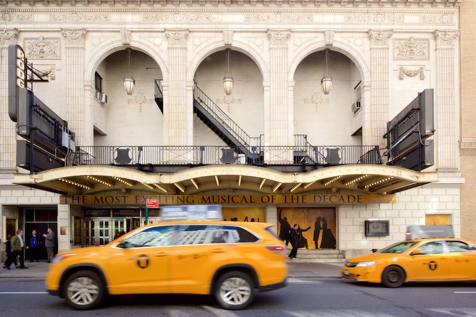 A theater with an ornate facade and a marquee reading "The Most Exciting Musical of the Decade".