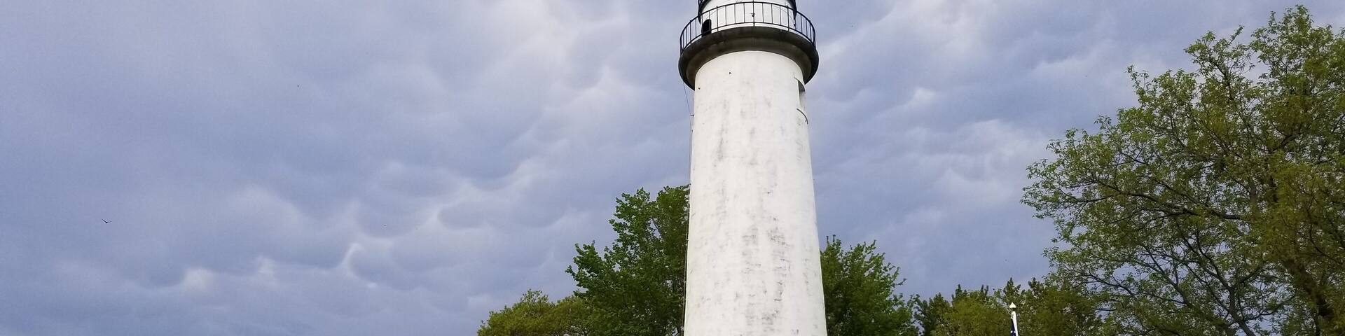 Un faro blanco con parte superior negra, una casa pequeña con terraza de madera y una bandera estadounidense.