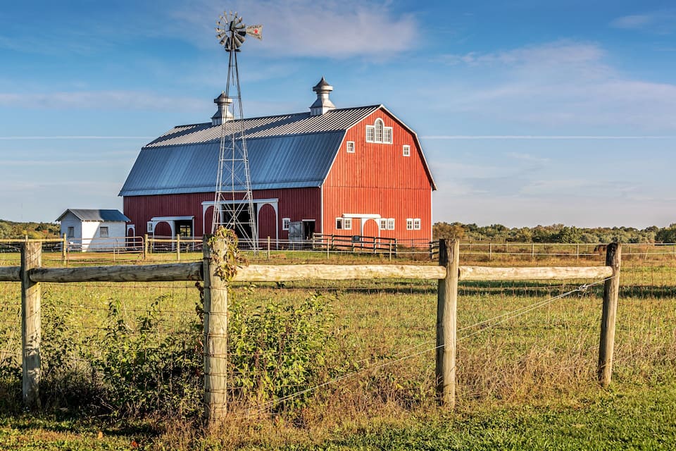 A red barn with a windmill, surrounded by a wooden fence and a grassy field.