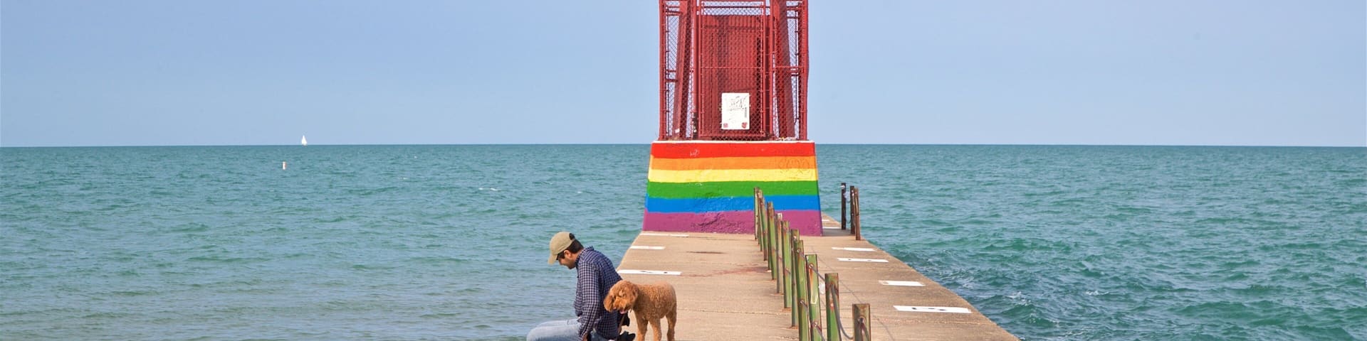 A lighthouse with a rainbow-colored base, a person and a dog on a pier.