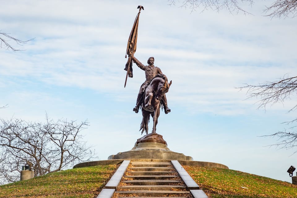 A statue of a mounted figure holding a flag, with a staircase leading up to it, surrounded by trees and a clear sky.