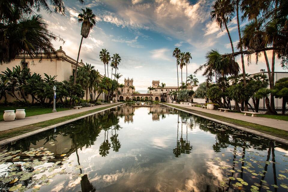 Un jardín simétrico con un estanque reflectante en el centro, palmeras y arquitectura clásica.