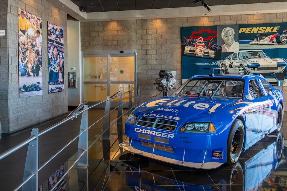 Une voiture de course bleue avec les inscriptions « Dodge » et « Charged », exposée dans un musée où l’on peut voir une affiche « Penske » et un panneau indiquant les toilettes.