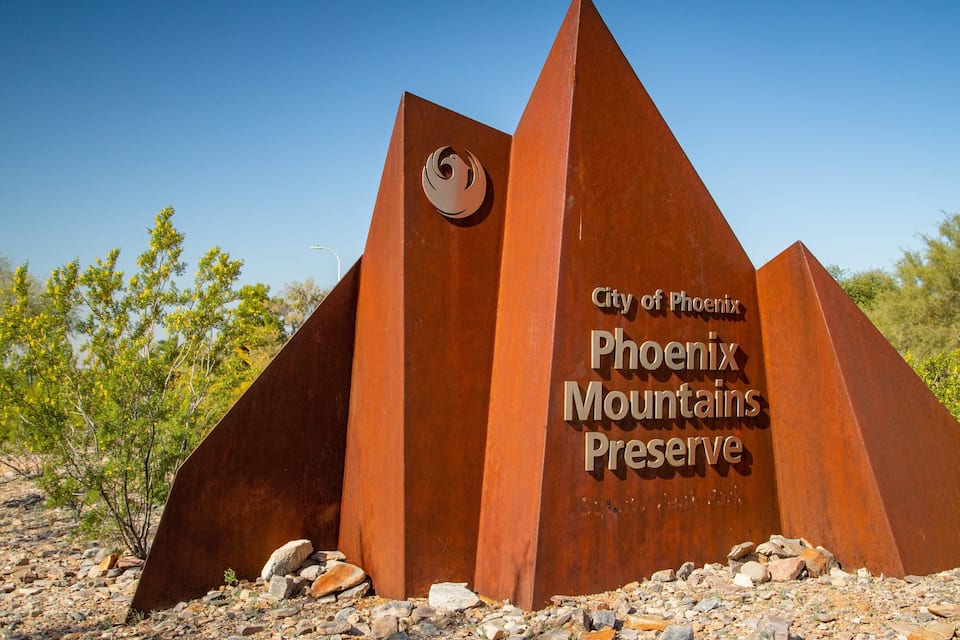 Ein großes, dreieckiges Metallschild mit dem Aufdruck 'City of Phoenix Phoenix Mountains Preserve' vor blauem Himmel.