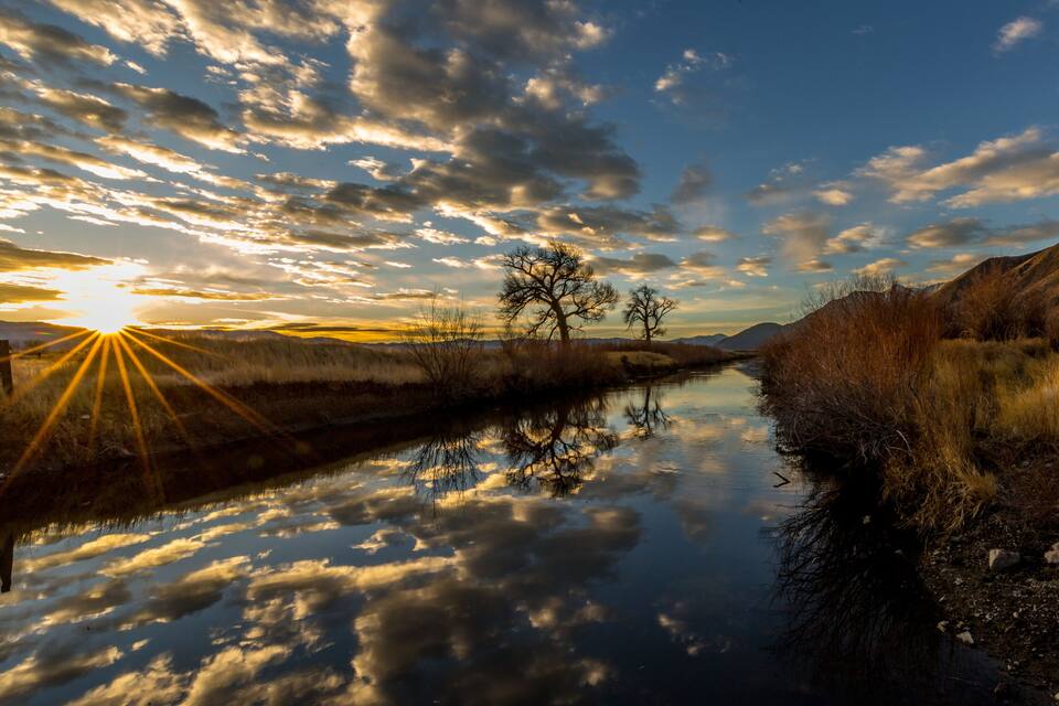 A river reflecting the sky with clouds, surrounded by dry grass and trees.