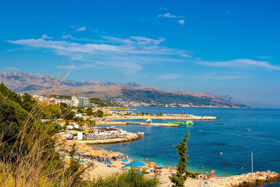 A coastal town with a beach, clear blue sea, mountains in the background, and a mix of buildings and greenery.