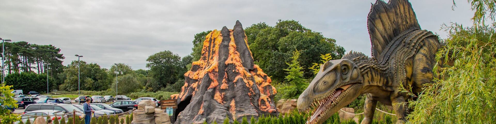 Un parque temático de dinosaurios con una gran estatua de dinosaurio, un volcán y un estacionamiento.