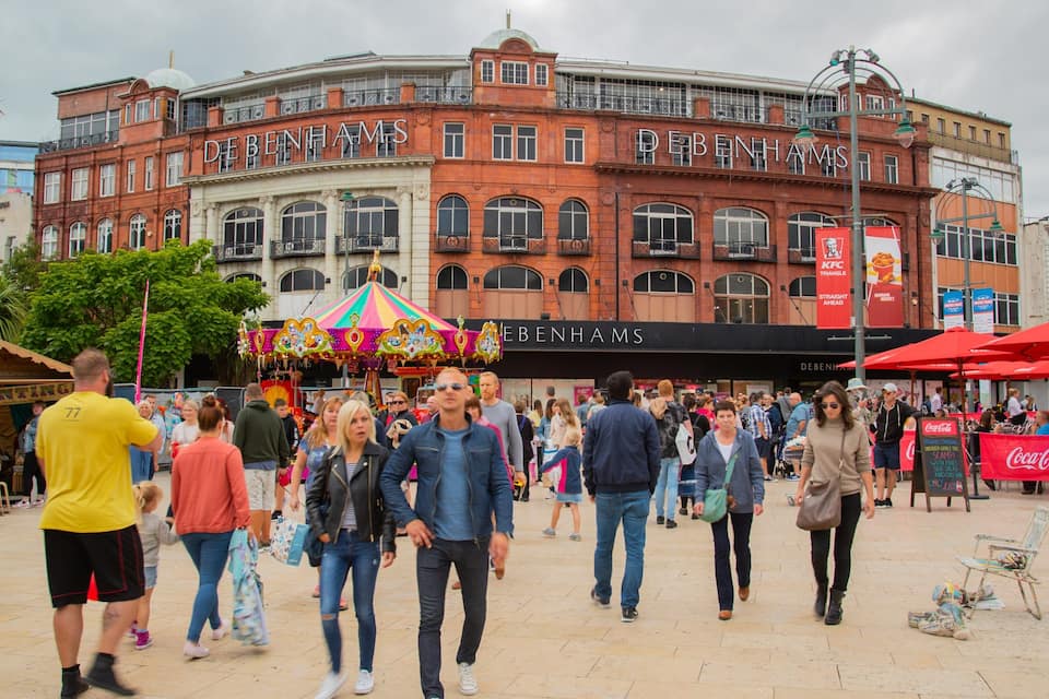 Una plaça pública animada amb un edifici vermell destacat que diu 'DEBENHAMS'.