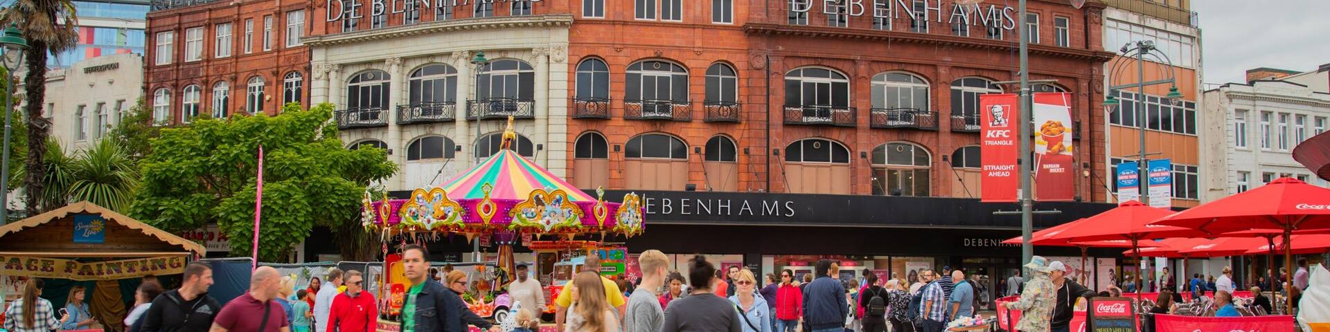 A busy public square with a carousel, people walking, and buildings with signs like 'DEBENHAMS' and 'KFC'.