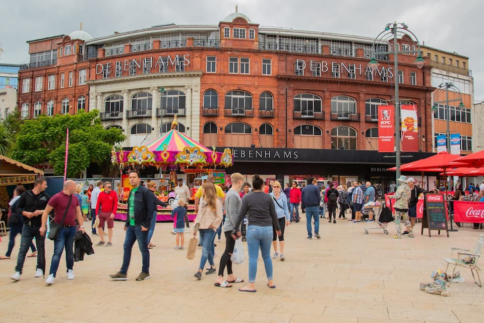 A busy public square with a carousel, people walking, and buildings with signs like 'DEBENHAMS' and 'KFC'.